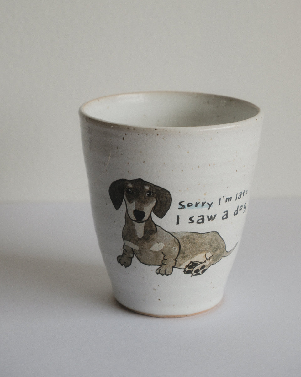 A handmade ceramic cup with a speckled off-white glaze, featuring a hand-drawn illustration of a dachshund and the text 'Sorry I'm late I saw a dog' printed on the front.
