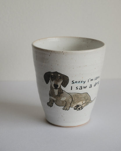 A handmade ceramic cup with a speckled off-white glaze, featuring a hand-drawn illustration of a dachshund and the text 'Sorry I'm late I saw a dog' printed on the front.