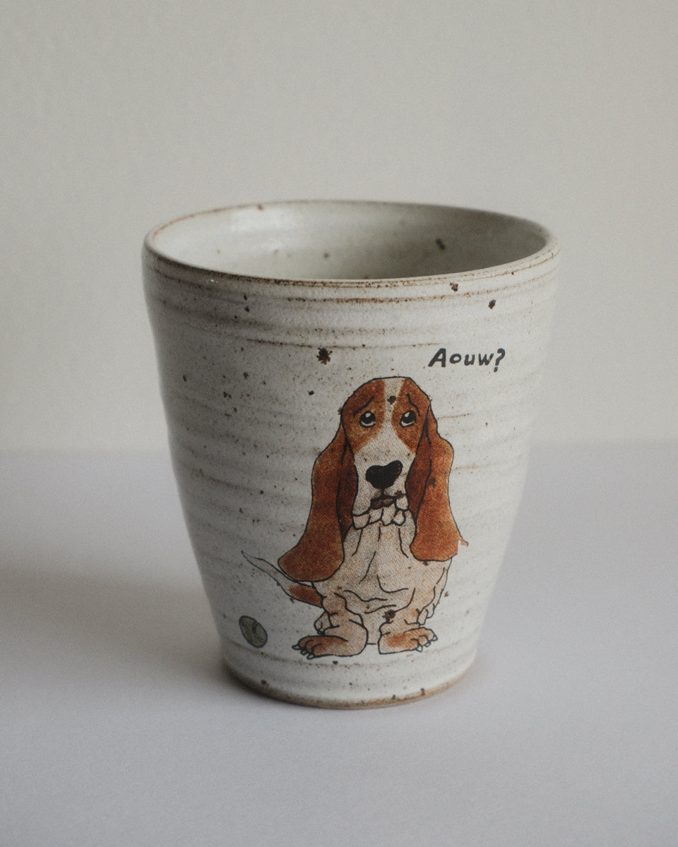 A handmade ceramic cup with a speckled off-white glaze, featuring a hand-painted brown and white Basset Hound illustration.
