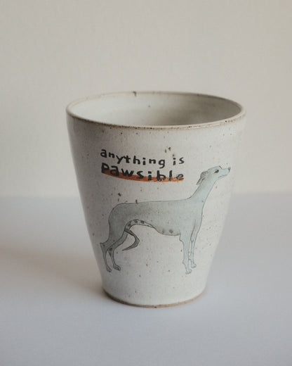 A handmade ceramic cup with a speckled off-white glaze, featuring a light blue-grey illustration of a greyhound and the text 'anything is Pawsible' on the front.