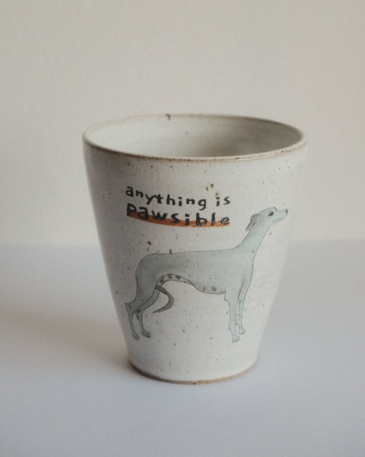A handmade ceramic cup with a speckled off-white glaze, featuring a light blue-grey illustration of a greyhound and the text 'anything is Pawsible' on the front.