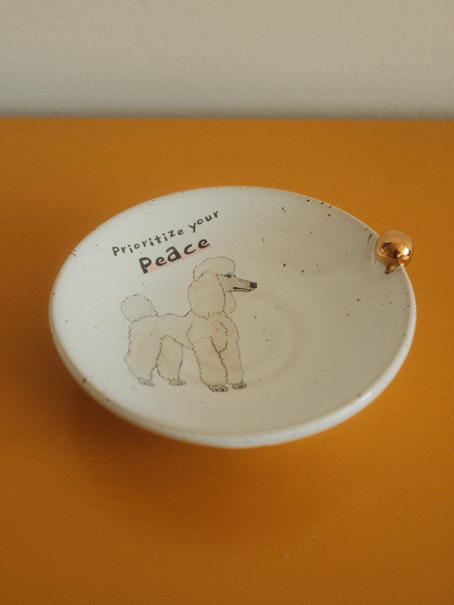 A small round white ceramic dish with a gold-tone accent on the rim, featuring a hand-drawn poodle and the text 'Prioritize your Peace' on the inside, set against a solid orange background.