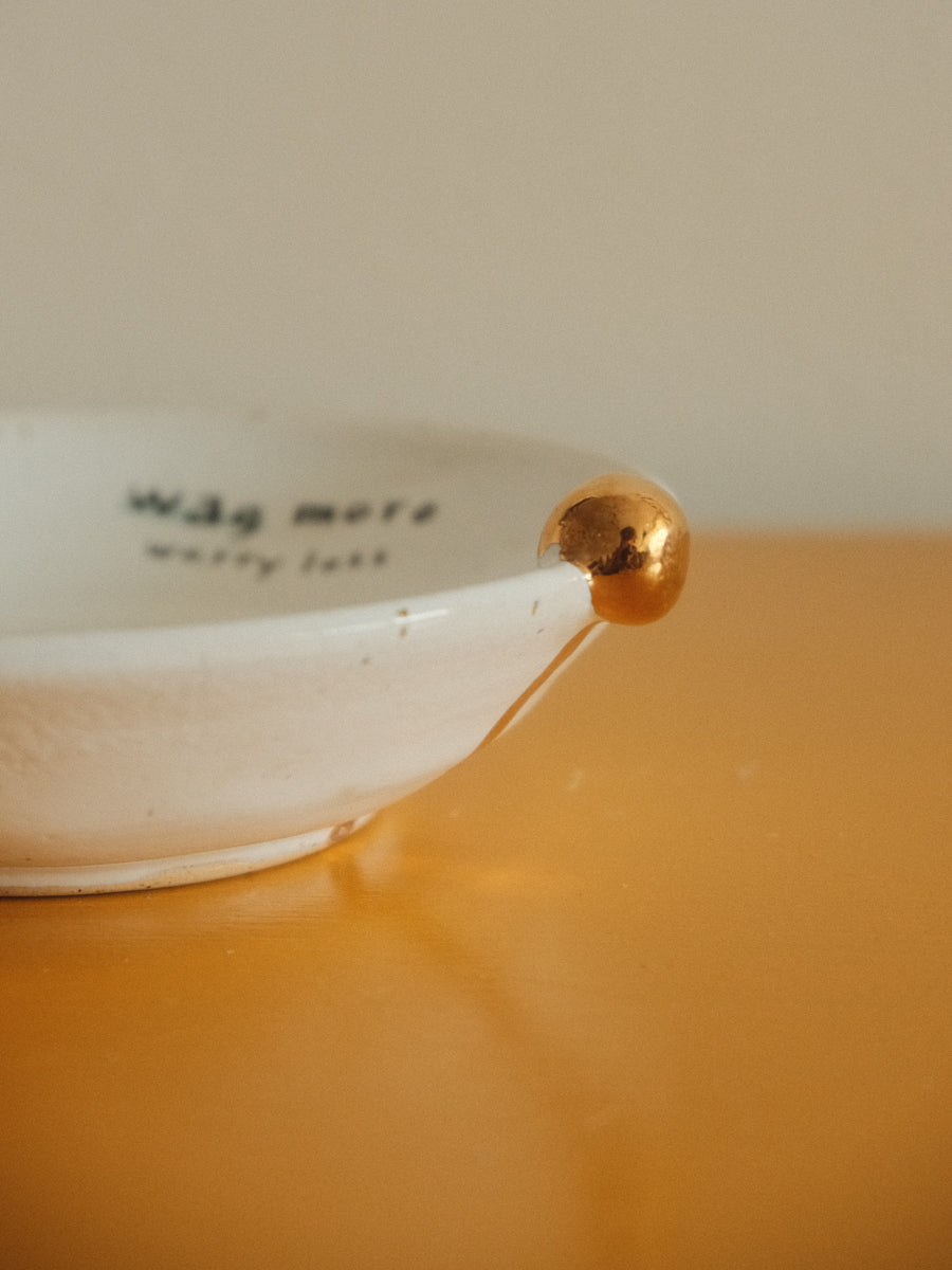 A close-up of a white ceramic bowl with a metallic gold accent on its rim, resting on a warm orange surface against a beige background. The inside of the bowl has black text that reads 'wag more warry leaf'.