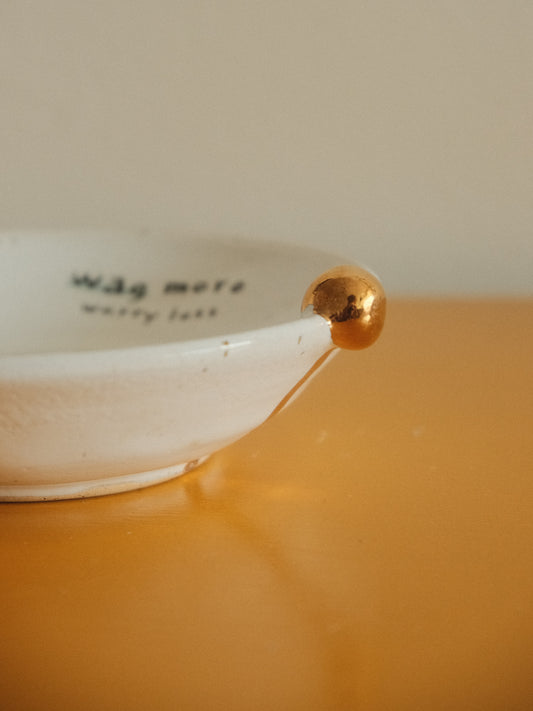 A close-up of a white ceramic bowl with a metallic gold accent on its rim, resting on a warm orange surface against a beige background. The inside of the bowl has black text that reads 'wag more warry leaf'.